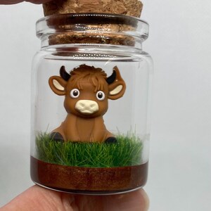 K&ouml;nnte beinhalten: Ein kleines Glasgef&auml;&szlig; mit einem Korkverschluss enth&auml;lt eine braune Tonfigur eines Bullen mit gro&szlig;en Augen. Der Bulle sitzt auf einem Bett aus gr&uuml;nem Gras mit einer braunen Basis.