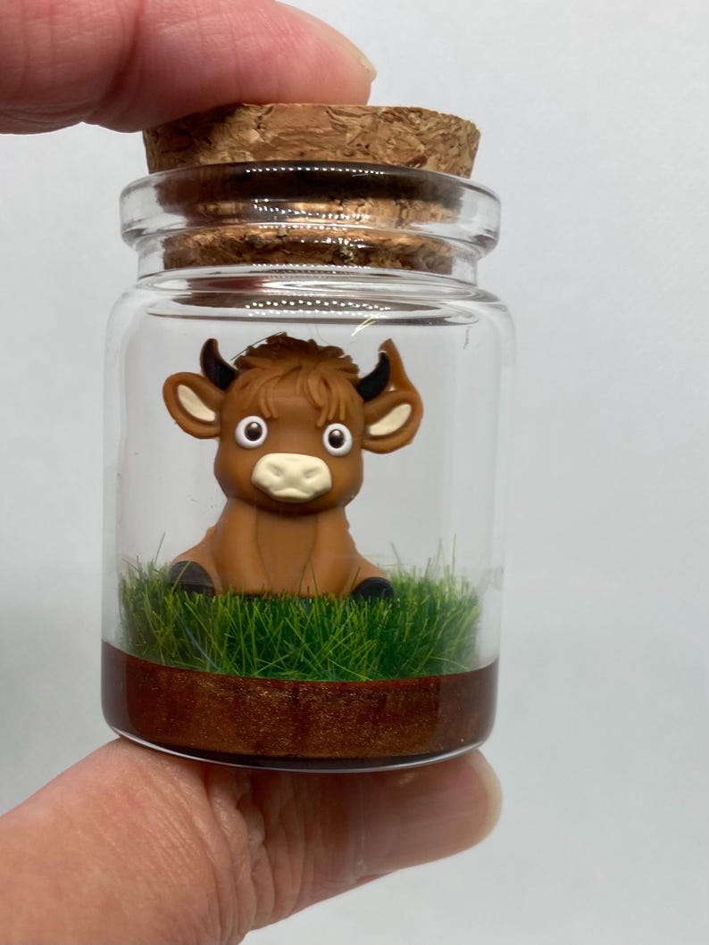 K&ouml;nnte beinhalten: Ein kleines Glasgef&auml;&szlig; mit einem Korkverschluss enth&auml;lt eine braune Tonfigur eines Bullen mit gro&szlig;en Augen. Der Bulle sitzt auf einem Bett aus gr&uuml;nem Gras mit einer braunen Basis.
