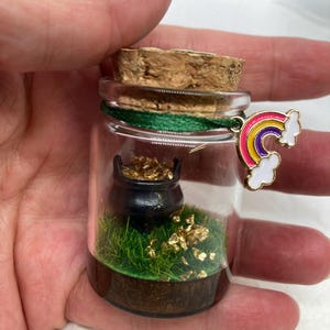 May include: A small glass jar with a cork lid contains a miniature pot of gold, green grass, and gold glitter. A rainbow charm hangs from the jar.
