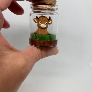 K&ouml;nnte beinhalten: Ein kleines Glasgef&auml;&szlig; mit einem Korkverschluss enth&auml;lt eine braune Miniatur-Stierfigur, die auf einem Bett aus gr&uuml;nem Gras sitzt. Das Gef&auml;&szlig; hat einen braunen Boden.