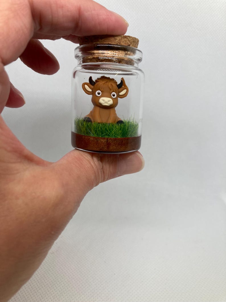 K&ouml;nnte beinhalten: Ein kleines Glasgef&auml;&szlig; mit einem Korkverschluss enth&auml;lt eine braune Miniatur-Stierfigur, die auf einem Bett aus gr&uuml;nem Gras sitzt. Das Gef&auml;&szlig; hat einen braunen Boden.