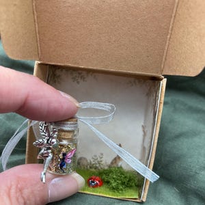 May include: A miniature glass bottle with a cork stopper containing gold glitter and a small fairy figure, adorned with a silver metal fairy charm and a thin silver ribbon. The bottle is presented inside a small cardboard box with a printed woodland scene.
