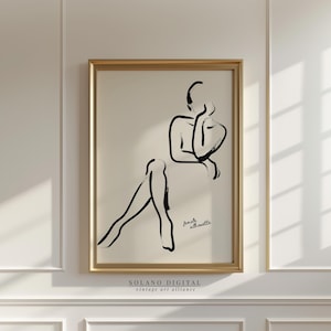 Może przedstawiać: Oprawiony wydruk z minimalistycznym czarnym tuszem przedstawiającym siedzącą postać. Grafika, oznaczona jako "female silhouette", jest umieszczona na kremowym tle w złotej ramie. Wydruk jest zamontowany na białej ścianie.