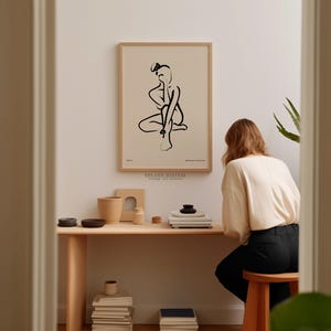 A woman is sitting at a wooden desk, surrounded by various objects such as books, bowls, and a plant, in a room with a framed abstract drawing hanging on the wall.