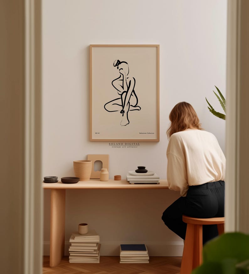 A woman is sitting at a wooden desk, surrounded by various objects such as books, bowls, and a plant, in a room with a framed abstract drawing hanging on the wall.