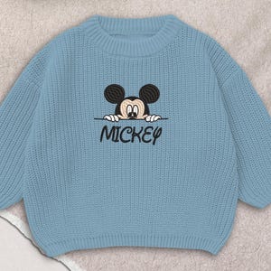 Puede incluir: Un suéter de punto azul claro con un diseño bordado de Mickey Mouse. El diseño incluye la cara de Mickey y la palabra "MICKEY" en escritura negra. El suéter tiene cuello redondo y mangas largas.