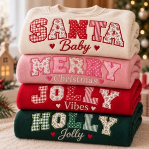 May include: A stack of four folded sweatshirts in cream, pink, red, and dark green. Each features large, patterned fabric letters spelling SANTA Baby, MERRY Christmas, JOLLY Vibes, and HOLLY Jolly, accented with small embroidered hearts.