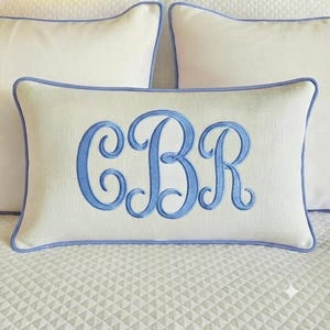 May include: A rectangular off-white throw pillow featuring a light blue embroidered monogram with the letters CBR in a cursive script font. The pillow is finished with a light blue piped edge trim that outlines the entire rectangular shape.