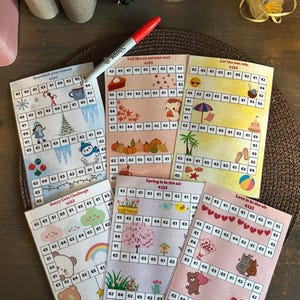 Op de afbeelding: Zes rechthoekige papieren spaarchallenge-trackers met rastervakjes voorzien van eurobedragen. De thema's omvatten winter, herfst, zomer, lente en liefde, met kleurrijke illustraties en een rode markeerstift die bovenop de kaarten ligt.