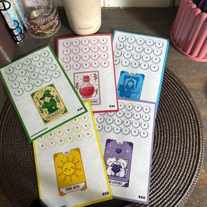 Op de afbeelding: Vijf rechthoekige papieren spaarchallenge-kaarten met gekleurde randen. Elke kaart bevat een illustratie in tarotstijl, waaronder De Magiër, De Zon, De Geliefden en een flesje, samen met rijen genummerde cirkels om spaardoelen bij te houden.