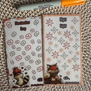 Op de afbeelding: Twee rechthoekige spaarchallenge-kaarten met een oranje permanente marker. De linkerkaart toont een wasbeer en de tekst Marshmellow Drop. De rechterkaart toont een vos, een lantaarn en de tekst Night Light. Beide kaarten bevatten de tekst 50.