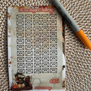 Op de afbeelding: Een rechthoekige spaarkaart met de titel 50 enveloppen challenge, voorzien van vijftig genummerde envelop-iconen, een illustratie van een wasbeer, de tekst 1275 en AGirlWithACatBudget, vergezeld door een grijze en oranje marker.