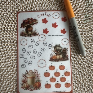 May include: A rectangular sticker sheet titled Cozy Fall featuring illustrations of a hedgehog with an umbrella, a raccoon by a campfire, a latte, and maple leaves. It includes circular tracking icons with euro symbols and an orange Sharpie marker.