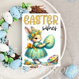 May include: A white bunny wearing a blue dress and a flower crown holds a blue and green Easter egg. The bunny is painting the egg with a paintbrush. The text "Easter wishes" is written in yellow letters on a white background.