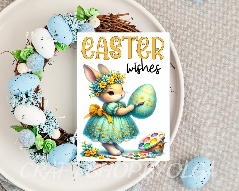 May include: A white bunny wearing a blue dress and a flower crown holds a blue and green Easter egg. The bunny is painting the egg with a paintbrush. The text "Easter wishes" is written in yellow letters on a white background.