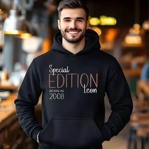 Könnte beinhalten: Schwarzer Kapuzenpullover mit Fronttasche und Kapuze. Der Hoodie hat den Text "Special EDITION Born in 2008 Leon" in weißer und brauner Schrift. Die Person auf dem Bild lächelt.