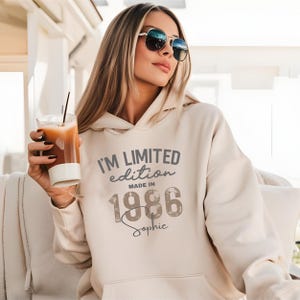 Könnte beinhalten: Ein beigefarbener Kapuzenpullover mit dem dunkelgrauen Text I'M LIMITED edition MADE IN 1986 und dem Namen Sophie in Schreibschrift darunter. Der Hoodie hat lange Ärmel, eine Kängurutasche und eine lockere Passform.