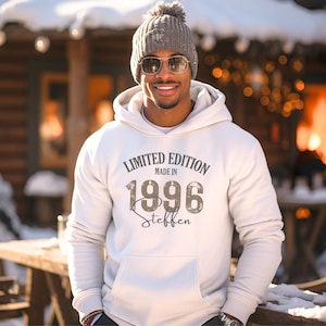 Könnte beinhalten: Ein cremefarbener Kapuzenpullover mit dem Text "LIMITED EDITION MADE IN 1996 Steffen" in einer dunkelgrauen, abgenutzten Schriftart. Der Hoodie verfügt über eine Kängurutasche, lange Ärmel mit Rippbündchen und eine passende Kapuze mit Kordelzug.