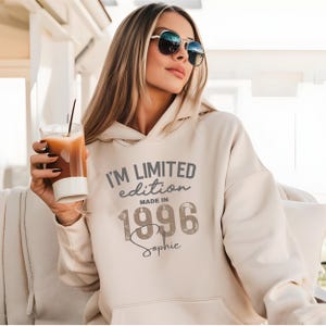 Könnte beinhalten: Ein beigefarbener Kapuzenpullover mit dem dunkelgrauen Text I'M LIMITED edition MADE IN 1996 und dem Namen Sophie in Schreibschrift. Das Kleidungsstück hat lange Ärmel, eine Kängurutasche auf der Vorderseite und eine Kapuze ohne Kordelzug.