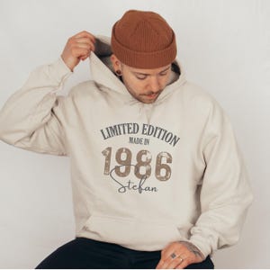 May include: A beige hoodie with a hood and the text "LIMITED EDITION MADE IN 1986 Stefan" in a distressed font. The person in the image is wearing a brown beanie.