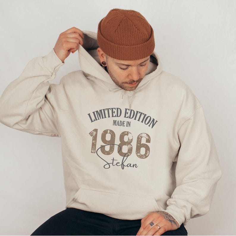 May include: A beige hoodie with a hood and the text "LIMITED EDITION MADE IN 1986 Stefan" in a distressed font. The person in the image is wearing a brown beanie.