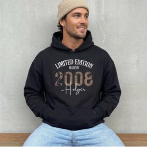 May include: Black hoodie with the text "LIMITED EDITION MADE IN 2008 Holger" in white and brown lettering. The hoodie has a front pocket and a drawstring hood. The person in the image is wearing a beige beanie and blue jeans.