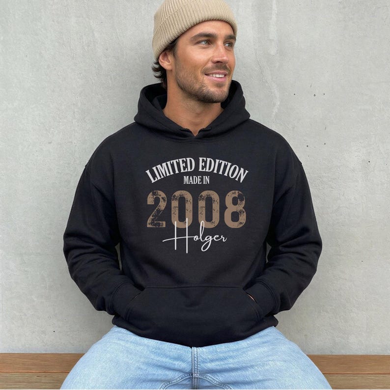May include: Black hoodie with the text "LIMITED EDITION MADE IN 2008 Holger" in white and brown lettering. The hoodie has a front pocket and a drawstring hood. The person in the image is wearing a beige beanie and blue jeans.