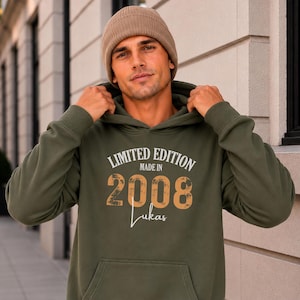 May include: An olive green hoodie with the text "LIMITED EDITION MADE IN 2008 Lukas" in white and orange. The hoodie has a front pocket and a drawstring hood. A person is wearing a brown beanie.