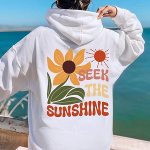K&ouml;nnte beinhalten: Wei&szlig;er Hoodie mit einem Sonnenblumen- und Sonnenmotiv und dem Text "Seek the Sunshine".