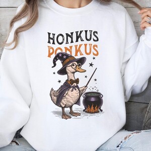 以下が含まれることがあります： 帽子をかぶって杖を持ったアヒルのグラフィックがプリントされた白いスウェットシャツ。アヒルの上に「Honkus Ponkus」という文字がプリントされています。
