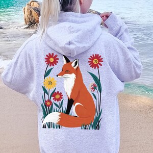 May include: Light gray hoodie with a fox and floral graphic on the back. The fox is orange, white, and black, surrounded by red, yellow, and green flowers. The word "Ash" is printed in the bottom right corner.