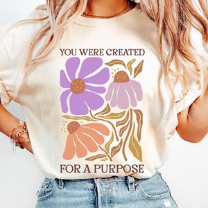 May include: A white t-shirt with the text "You were created for a purpose" printed on it. The text is in brown lettering and is surrounded by a floral design with purple, pink, and orange flowers.