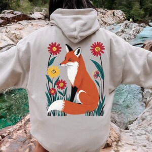 May include: Sand-colored hoodie featuring a vibrant illustration of a fox amidst colorful flowers. The fox is depicted in shades of orange, white, and black, surrounded by red and yellow floral accents. The hoodie has a drawstring hood and a kangaroo pocket.