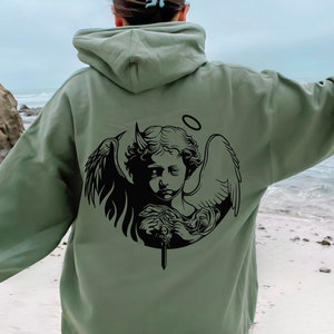 Może przedstawiać: Zielona bluza z kapturem w kolorze khaki z czarnym nadrukiem graficznym przedstawiającym anioła z rogami diabła trzymającego miecz. Anioł ma aureolę nad głową.