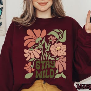 May include: A maroon sweatshirt featuring a floral design with the text "STAY WILD". The flowers are in shades of pink and peach with green leaves. The word "MAROON" is printed in the bottom right corner.