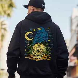 以下が含まれることがあります： 背景に三日月のある、頭蓋骨の上に止まっている青いカラスのグラフィックプリントが施された黒いフード付きスウェットシャツです。