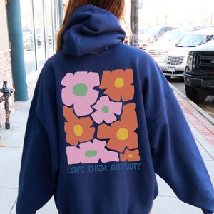 May include: A navy blue hooded sweatshirt featuring a large graphic on the back. The design consists of several stylized flowers in shades of pink, orange, and yellow with green centers. Below the floral graphic, the text reads LOVE THEM ANYWAY.