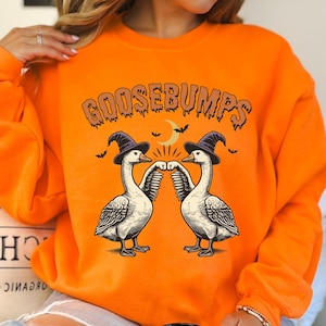 以下が含まれることがあります： オレンジ色のスウェットシャツに、魔女の帽子をかぶって拳を突き上げている2羽のガチョウのグラフィックが描かれています。ガチョウの上に「Goosebumps」という文字があります。