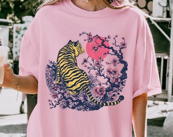 以下が含まれることがあります： ピンクのオーバーサイズTシャツに、黄色と黒の虎がピンクと赤の桜の木の前に座っているグラフィックプリントが施されています。グラフィックは日本のスタイルです。