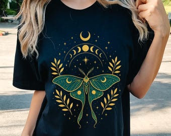 May include: Black t-shirt featuring a green and gold moth design with a crescent moon and phases of the moon. The design is surrounded by a wreath of leaves. The word "BLACK" is printed at the bottom of the shirt.