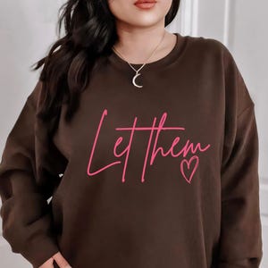 以下が含まれることがあります： ピンク色の「Let them」の文字とハートが特徴の、ダークチョコレートブラウンのスウェットシャツ。「Dark Chocolate」の文字が下部にプリントされています。長袖とクルーネックのデザインです。