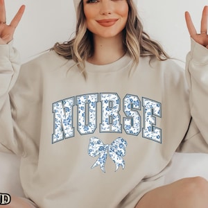 Könnte beinhalten: Ein beiger Pullover mit dem Wort "NURSE" in blau mit einem Blumenmuster bedruckt. Das Wort ist in einer College-Schriftart mit einer blauen Schleife darunter.