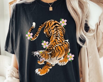 以下が含まれることがあります： ピンクの花がついた虎のグラフィックが描かれた黒いTシャツ。虎はオレンジと黒で、背景は白。画像の下部に「BLACK」という文字があります。