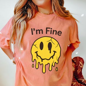 以下が含まれることがあります： テラコッタ色のTシャツ。上部に「I'm Fine」の文字、その下に溶けた黄色のスマイリーフェイスのグラフィック。Tシャツの下部には太字で「TERRACOTTA」の文字がプリントされています。