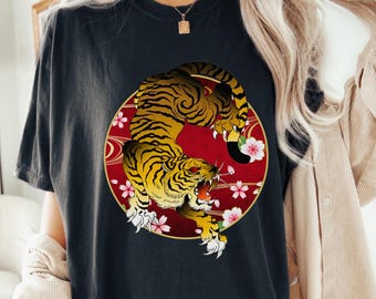 以下が含まれることがあります： 赤色の円の中に、黄色と黒の虎が描かれた黒いTシャツ。ピンクの桜がアクセントになっています。「BLACK」の文字が下にプリントされています。カジュアルな装いにぴったりの、おしゃれなグラフィックTシャツです。