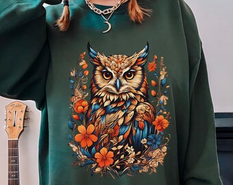 May include: Forest green sweatshirt featuring a detailed owl illustration surrounded by orange, blue, and yellow flowers. The owl has intricate feather patterns in shades of brown, blue, and orange. The text "FOREST GREEN" is at the bottom left.