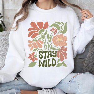 May include: White sweatshirt featuring a floral design in orange, peach, and green. The words "STAY WILD" are printed in green. The sweatshirt has a crew neck.