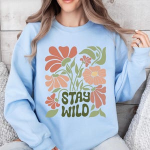 以下が含まれることがあります： オレンジ、ピーチ、グリーン系の花柄がプリントされたライトブルーのスウェットシャツ。「STAY WILD」の文字は緑色でプリントされています。画像の右下隅には「LIGHT BLUE」の文字があります。