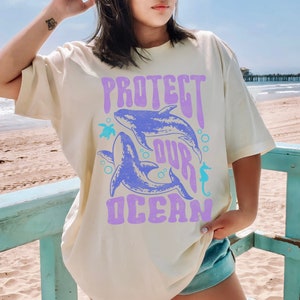 Pu&ograve; includere: Una maglietta beige chiaro con una stampa grafica di due balene blu e il testo "PROTECT OUR OCEAN" in viola. La grafica include anche un cavalluccio marino e una tartaruga marina.