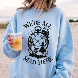 Puede incluir: Sudadera azul claro con un gráfico negro que presenta un reloj, personajes de Alicia en el País de las Maravillas y el texto "WE'RE ALL MAD HERE". Las palabras "LIGHT BLUE" están en la parte inferior de la sudadera.